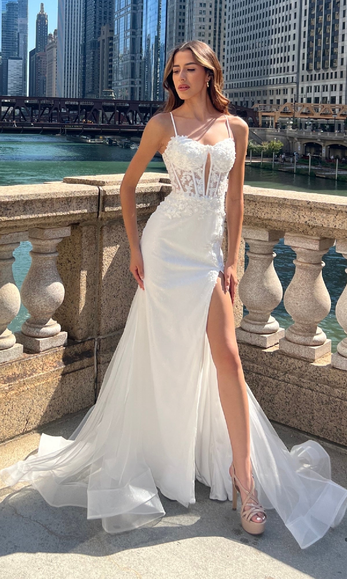 Woman in an Amelia Couture 7064 Long Mermaid Prom Dress with a sheer bodice and high slit stands on a stone terrace by a river, exuding elegance like a floral lace gown amid tall urban buildings.